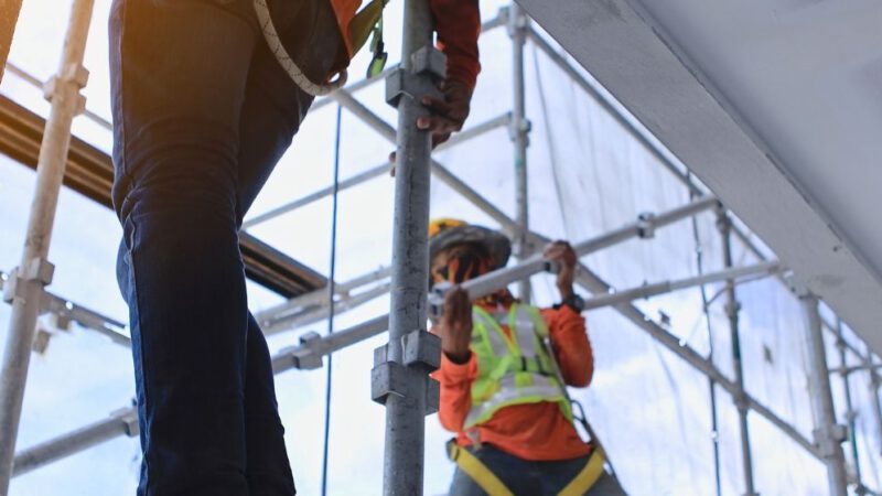 Zwei Menschen sind auf einem Baustellengerüst gesichert, stehen auf dem Gerüst, es ist von der Gebäudeinnenseite aus fotografiert